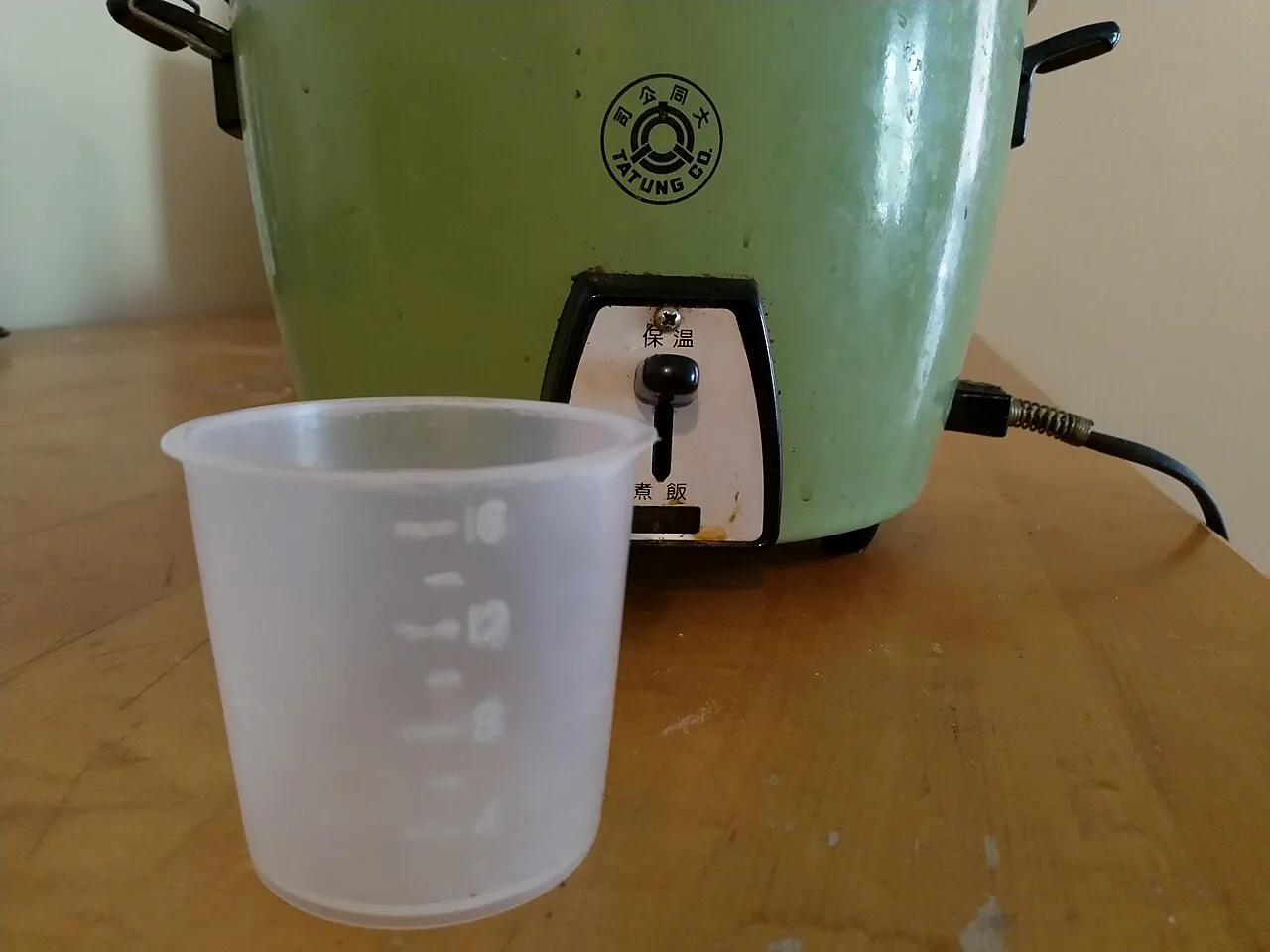 Gobelet doseur en plastique de 180 millilitres devant un cuiseur à riz